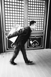 A black-and-white image shows a person in casual attire walking while carrying a large box on their back, capturing a moment of travail in Pékin. The background features ornate doors with large square and rectangular patterns.