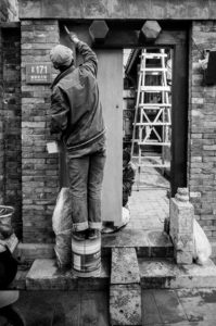 A person standing on a paint can paints the top of a doorway in a brick structure, reminiscent of scenes from Pékin. Nearby, another person crouches, working at ground level. A ladder and various materials are visible around them, indicating the site’s bustling travail and renovation efforts.