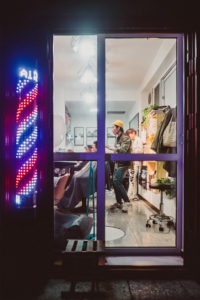 View inside a barbershop through a large glass door lit by a neon barber pole sign, reminiscent of the vibrant nights in Pékin. Several barbers are attending to their clients. The shop's interior features bright lighting, modern decor, and various grooming tools on countertops.