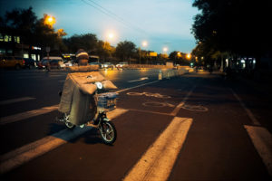 A person rides a small electric bike at dusk, bundled in a large, padded coat and scarf. The bike has a basket attached to the front. The scene is set on a city street in Pékin with bike lanes, streetlights, and cars in the background.