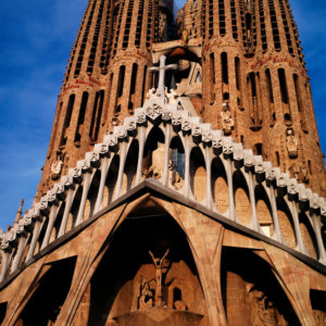 The image shows the Nativity facade of the Sagrada Família, a large Roman Catholic basilica in Barcelone, Spain. The facade features ornate architectural details, including sculpted figures and a prominent cross at the center, set against a clear blue sky.