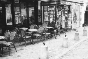 A black-and-white photo of a street scene with a man, Alain, sitting alone at an outdoor café table. The café, named Montmartre Tabacs, has empty woven chairs and tables lined up. Alain is wearing sunglasses and appears contemplative. Nearby there are postcard stands.