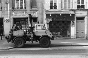 Black and white photo of a rugged, gros utility truck parked on the street in front of two old shops. One shop sign reads "AU VIEUX DOCUMENT" and the other reads "Tapissier Ensemblier." The camion has large tires and equipment mounted on its rear.