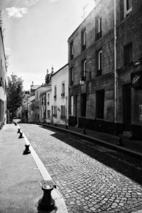 A black and white photo of a narrow, cobblestone street in Butte-aux-Cailles, flanked by tall buildings. Light casts dramatic shadows. Street lamps and bollards line the sidewalk. Trees and a dome-topped building are visible in the background under a partly cloudy sky, evoking Paris confiné.