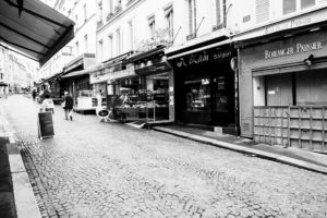 A black-and-white photo of a cobblestone street in Paris confiné. Shops line the right side, including a boulanger-pâtissier and VN Saigon restaurant. A person walks away in the distance, and part of a café canopy is visible on the left. The quiet Rue Mouffetard appears deserted.
