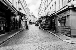 Black and white photograph of a narrow cobblestone street lined with various storefronts and awnings. This empty stretch captures the old-world charm of Rue Mouffetard, with its mix of architectural features in a scene reminiscent of Paris confiné.
