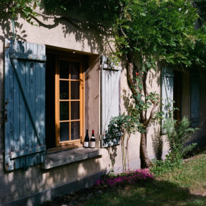 A rustic house wall in La Celle-les-Bordes features three blue wooden shutters, two of which are closed. The middle window is open, with two wine bottles placed on its sill. Plants and flowers grow around the base of the walls, and sunlight casts dappled shadows across the scene.