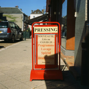A red sandwich board on a street reads "PRESSING" and advertises a special cleaning and disinfection program related to COVID-19, following WHO guidelines. Titled "Sans Titre," the sign is placed in front of a shop on a sunny day with buildings and a car visible in the background.