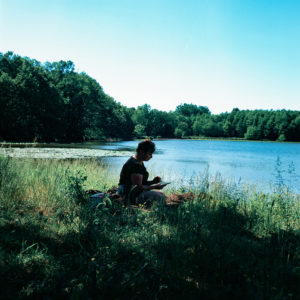 A person with short hair sits in the shade by a tranquil lake, surrounded by tall grass and trees. They are looking down, possibly reading or writing, with the sunlight casting a soft glow over the scene—an image that could be titled Sans Titre for its serene ambiguity.