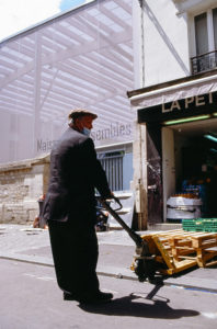 A man in a suit and flat cap uses a pallet jack to move wooden pallets on a narrow street. He stands next to an open entrance labeled "LAPET." The background shows part of a building with a large banner displaying the text "Maison assemblée," reminiscent of the vibrant Marché d'Aligre nearby.