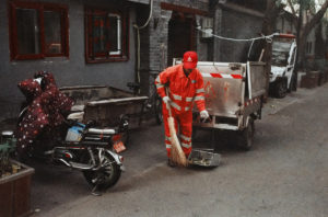 A street cleaner in a bright orange uniform sweeps the ground on a narrow hutong with a traditional-style building in the background. The cleaner has a broom and dustpan. Nearby, a motorcycle and a small utility vehicle are parked beside the road, giving an authentic glimpse of Pékin, Chine.