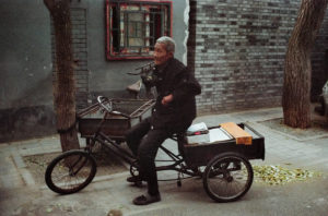 An elderly person sits on a tricycle cart on a narrow hutong in Pékin. They are wearing a dark jacket and have gray hair. Behind them is a brick wall with a window framed in red. The ground is scattered with leaves.