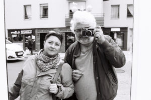 A black and white photo, titled *Sans Titre*, features a man and woman outdoors. The woman on the left wears a jacket and scarf, smiling slightly. The man on the right, with glasses and gray hair and beard, holds a camera up to his eye, capturing the moment. Urban background with buildings and cars.