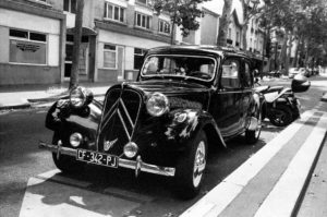 A black and white photo captures a vintage car, identified as a Traction Avant, parked on a city street. The car, with license plate CF-342-PJ, features a distinct grille and rounded headlights. A modern motorcycle is parked next to it, with buildings lining the background street scene.