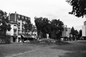 A black-and-white photo of a quiet town square showcases a circular roundabout. A statue stands in the center surrounded by trees and small flowerbeds. Buildings with shops and balconies line the background, and a car is parked along the street, reminiscent of a quaint place along the Danube.