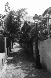 A black-and-white photograph of a narrow, cobblestone pathway in the Quartier de la Mouzaïa, lined with trees and a vintage streetlamp on the right. The pathway is flanked by fences and walls, leading uphill towards a taller building in the distance.