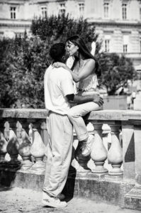 A black and white photo of Les amoureux du parc sharing an intimate moment. The woman is sitting on a stone railing, holding the man's shoulder, while he stands between her legs, looking up at her. They are outdoors with a building and trees in the background.