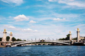 A view of the Pont Alexandre III bridge in Paris, France, one of the most splendid French bridges spanning the Seine River. The ornate bridge features golden statues and decorative street lamps. The water in the foreground reflects the bridge, and the sky above is blue with scattered clouds.