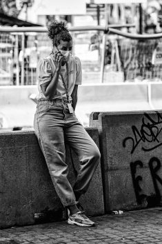 A woman with curly hair, wearing a mask, t-shirt, high-waisted jeans, and sneakers, leans against a concrete barrier while talking on her phone. Against the backdrop of Barcelone's bustling Eixample district construction site with fencing and barriers. The photo is in black and white.