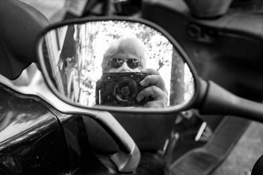 A monochrome photo capturing the reflection of a person wearing sunglasses, holding a camera, and taking a picture through the side mirror of a motorcycle. The background shows blurred trees and part of a building in Barcelone's Eixample district.