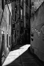 A narrow, deserted alleyway in Barcelone's Barri Gòtic quarter with tall buildings on either side. Graffiti decorates the right wall and a curved iron lamp hangs on the left. Light and shadows create a stark contrast on the cobblestone path. The scene is captured in black and white.