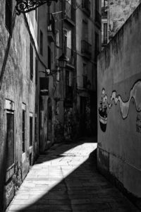 A narrow, deserted alleyway in Barcelone's Barri Gòtic quarter with tall buildings on either side. Graffiti decorates the right wall and a curved iron lamp hangs on the left. Light and shadows create a stark contrast on the cobblestone path. The scene is captured in black and white.
