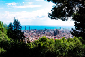 A panoramic view of Barcelone from Turó de la Rovira, framed by trees. The cityscape includes the Sagrada Familia and two high-rise towers. The Mediterranean Sea is visible in the background under a partly cloudy sky.