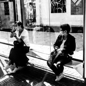 A black and white image of two individuals wearing masks and sitting on a bench at a bus stop in Gràcia, Barcelone. The person on the left is adjusting their mask, while the one on the right is looking away. The background includes posters and graffiti on the bus stop shelter.