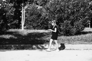 A woman in a short, dark dress walks on a paved path beside bushes and trees in the Parc de la Ciutadella, Barcelone. She is looking at her phone and has her hair tied back. The scene is in black and white, with shadows from the foliage cast on the ground.