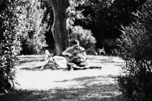A person sits on a blanket on the grass, playing a guitar in Parc de la Ciutadella, surrounded by trees and bushes. Another person is lying down nearby, enjoying the sunny day. The scene is serene and relaxed. The image is in black and white.
