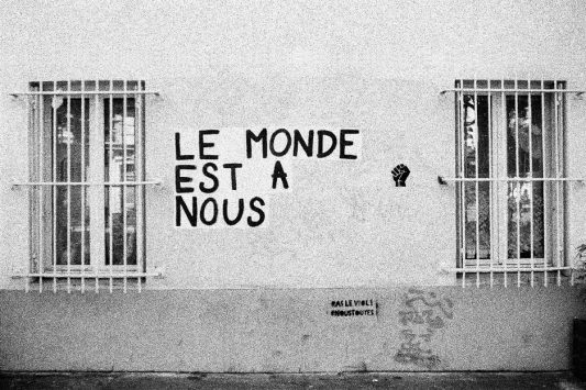 2020-07-31-Minox-GTE-35mm-Kodak-TMAX3200-minus-1-stop_20_MOD_BW_FRAME_resize Black and white image of a wall with two barred windows. Between the windows, graffiti reads "LE MONDE EST A NOUS" ("THE WORLD IS OURS") in French. A small drawing of a raised fist is next to the text. Smaller graffiti below reads "#ALELMNL #NOUSONEST" accompanied by "Sant Titre.