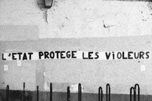 A black and white photograph of a wall with French graffiti that reads, "L'ÉTAT PROTÈGE LES VIOLEURS" in capital letters. The surroundings appear slightly worn, and the text is painted in a bold, stark style.