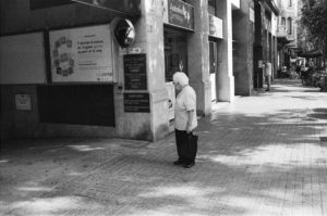 A black and white image of an elderly person with white hair standing alone on a city sidewalk in the Eixample district of Barcelone, holding a shopping bag. They are facing a wall with various signs and posters, and nearby there are buildings and a few pedestrians in the distance.