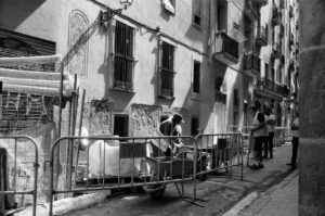 Black and white image of a street scene in Barcelona's Raval neighborhood with construction work in progress. Scaffolding and barricades line the sidewalk, while workers engage in restoration activities on a building. Pedestrians stand nearby; some look on, while others pass by.