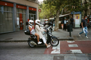 Two people, both wearing helmets, ride a black scooter at a crosswalk in Raval. The passenger wears a pink floral top, and the driver is dressed in a long white coat. They are stopped in front of a Santander bank, with pedestrians and the bustling street scene of Barcelone in the background.