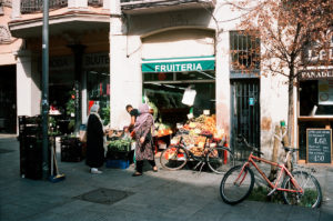 Exterior of a small storefront labeled "Fruiteria" in Raval, Barcelone, with fresh produce displayed outside. Two people in patterned clothing shop at the stand. The scene includes parked bicycles, a street vendor, and a neighboring shop sign displaying prices for café con leche and buns.