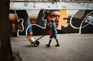 A person wearing a blue shirt, blue cap, and face mask is pushing a hand truck with a box on it along a sidewalk in the Raval neighborhood of Barcelona. The background features a mural with various abstract shapes and faces. A large tree is partly visible on the left side of the image.
