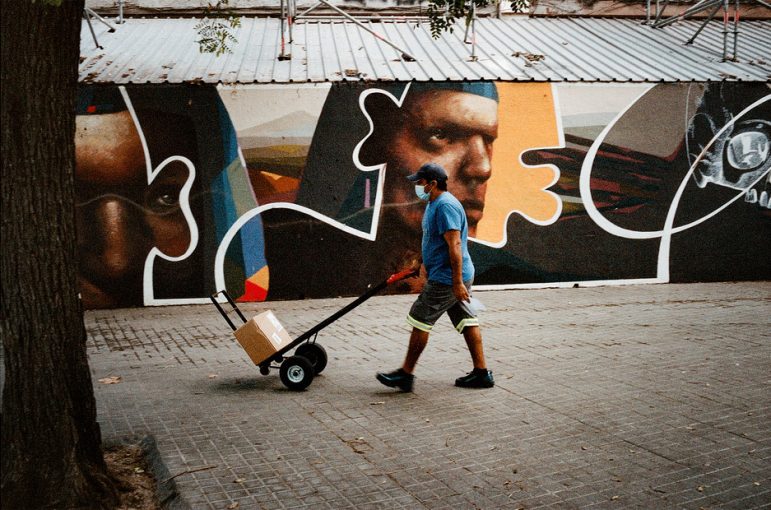 A person wearing a blue shirt, blue cap, and face mask is pushing a hand truck with a box on it along a sidewalk in the Raval neighborhood of Barcelona. The background features a mural with various abstract shapes and faces. A large tree is partly visible on the left side of the image.