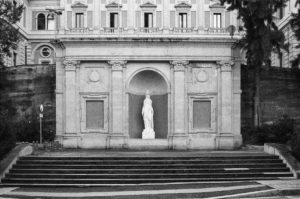 Black and white image of a classical building facade in Barcelone with a statue of a nude figure in the central archway. The building features ornate architectural details and is flanked by trees. Stone steps lead up to the entrance, creating a serene, timeless feel reminiscent of Poble Sec's charm.