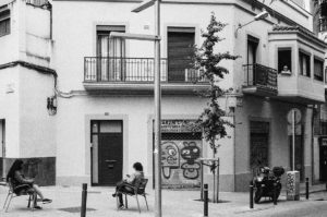 Black and white photo of a street corner in Barcelone's Poble Sec neighborhood: two people sit on chairs facing each other, a graffiti-covered storefront in the background. A person sits on a balcony above, while another individual leans on a moped next to a wall adorned with street art.