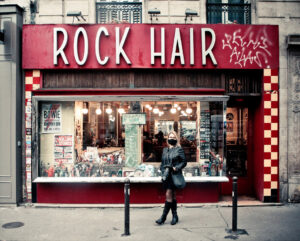 A person dressed in black stands outside "Rock Hair," a store with a red facade and checkered yellow accents, on a city street. The storefront window displays various posters and memorabilia, and lights illuminate the interior. Graffiti is visible on the signage.