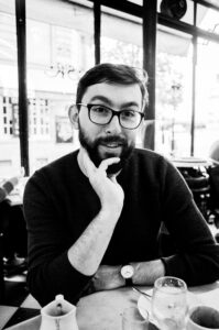 Black and white photograph of David Rothan, a bearded man with glasses, wearing a sweater and a watch, seated at a table in a café. His chin rests on his right hand, and there are drinks and a small pitcher on the table. Sunlight filters through the windows behind him.
