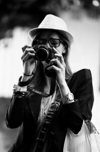 Black and white photograph of a femme photographe wearing a hat and glasses, holding a camera up to their eye, seemingly taking a photo. The individual has a watch and several wristbands on their left wrist, and wears a bag over their shoulder.