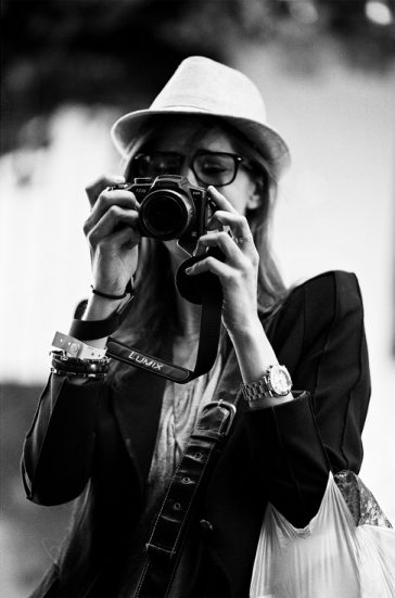 2011-05-21-Canon-EOS-1V-Kodak-TMAX400_F00000018_MOD_BW_FRAME_resize Black and white photograph of a femme photographe wearing a hat and glasses, holding a camera up to their eye, seemingly taking a photo. The individual has a watch and several wristbands on their left wrist, and wears a bag over their shoulder.