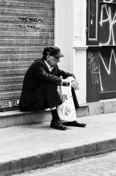 2011-05-21-Canon-EOS-1V-Kodak-TMAX400_F00000032_MOD_BW_FRAME_resize Black and white image of an older man named Marc Emile sitting on a step in a city. He wears a cap, a dark jacket, pants, and black shoes, holding a plastic shopping bag. He sits in front of a closed, graffiti-covered metal shutter and wall. He appears contemplative or tired.