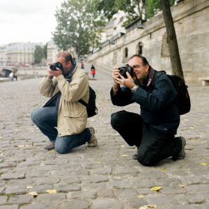 Two men, Fabrice Mercier and Helder Vinagre, kneel on cobblestone near a river, focused on taking photos with their cameras. One wears a beige jacket, blue jeans, and glasses, while the other sports a black jacket and pants. Trees and buildings line the background, adding a picturesque urban setting.