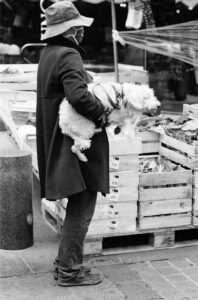 A person wearing a dark coat, hat, and mask stands next to wooden crates in the bustling Place du Marché, holding a small dog that is looking over the crates. The photo is in black and white.