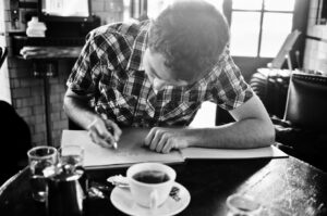 Black and white image of Grégoire Brossard with short hair wearing a plaid shirt, sitting at a table in a cafe. They are writing or drawing a dédicace in a large open book. On the table are a cup of coffee, a glass of water, and a couple of other items.