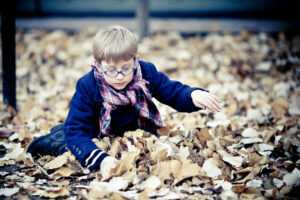 Le petit Jean-François, a young boy with blond hair and blue glasses, is sitting on the ground amidst a thick layer of fallen autumn leaves. He is wearing a dark jacket and a plaid scarf. The boy appears to be playing or exploring the leaves intently.