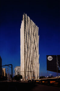 A modern skyscraper with a unique, crisscrossed exterior design stands tall against a clear blue sky. The structure, known as Torre Telefónica in Barcelona, is primarily composed of glass and metal. On the right, a dark flag with the word "nat" is visible, as well as part of the street below.